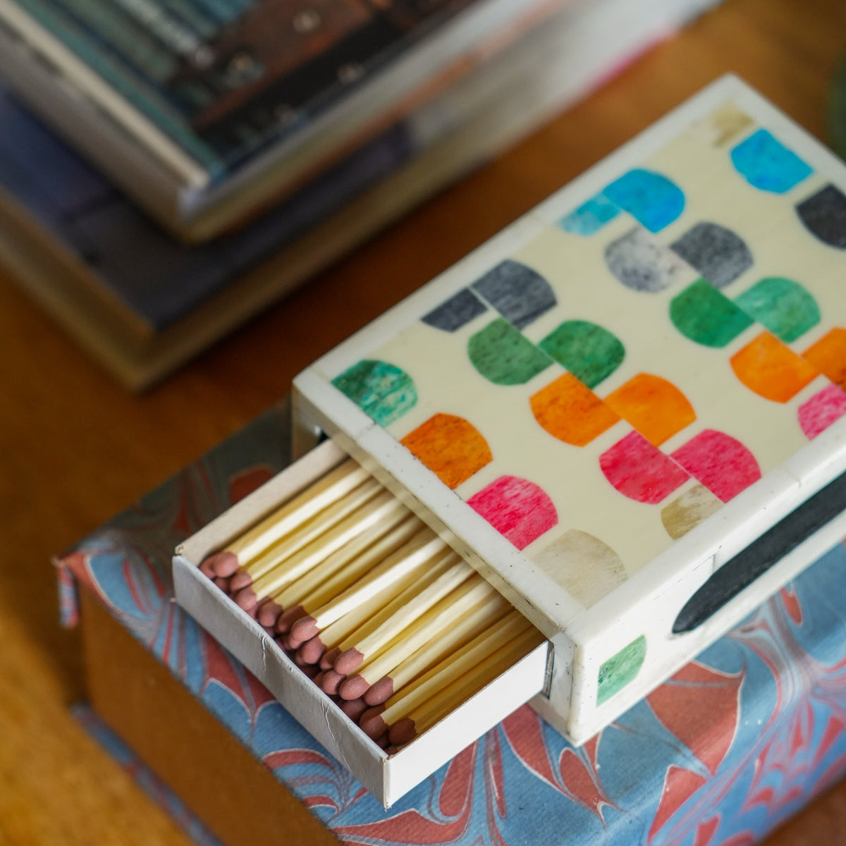 A decorative matchbox with a colorful geometric pattern sits open on top of a marbled box, showing wooden matches with pink tips, on a wooden surface.