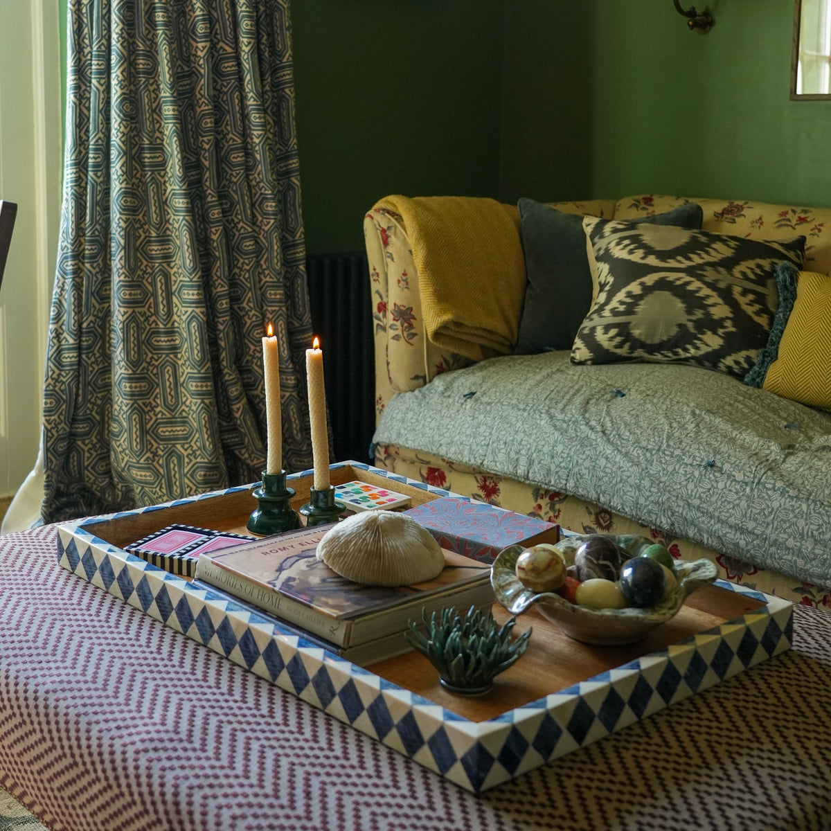 A styled interior scene with a patterned green wall, matching curtains, a floral sofa, candles, books, and a decorative tray on a table in a living room setting.