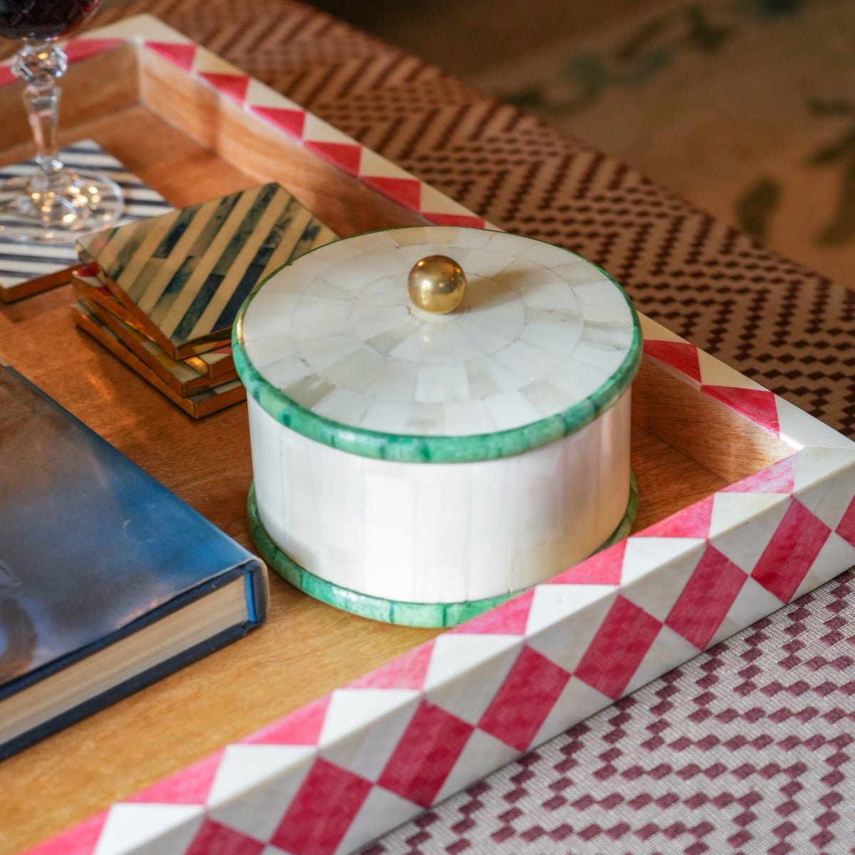 A round lidded box with a white inlay top and green trim sits on a geometric tray with pink and white patterned edges, styled on a wooden table with a book, coaster, and wine glass nearby.
