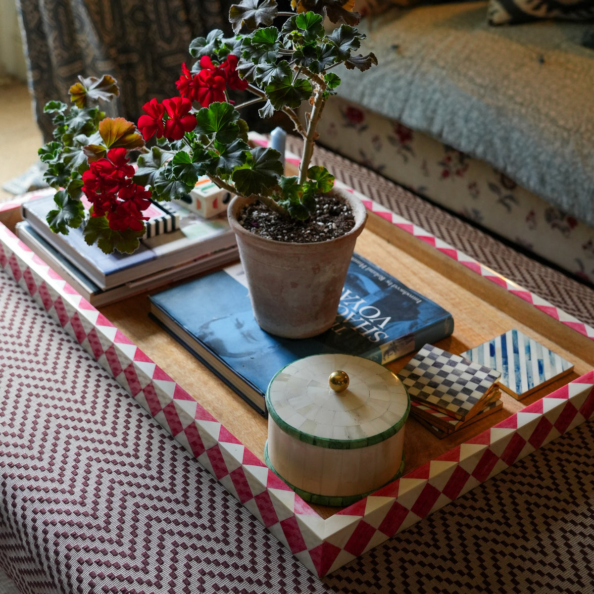 A styled living room scene showing a wooden tray with a pink and white geometric patterned border, holding a potted red flowering plant, books, a small round wooden box with a gold knob, and folded textiles. A sofa and curtains are visible in the background.