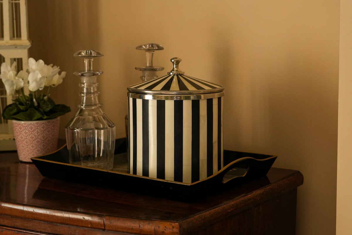 A black and white vertically striped decorative jar with a matching lid sits on a black serving tray on a dark wooden chest, next to clear glass decanters, with a beige wall and framed art in the background.