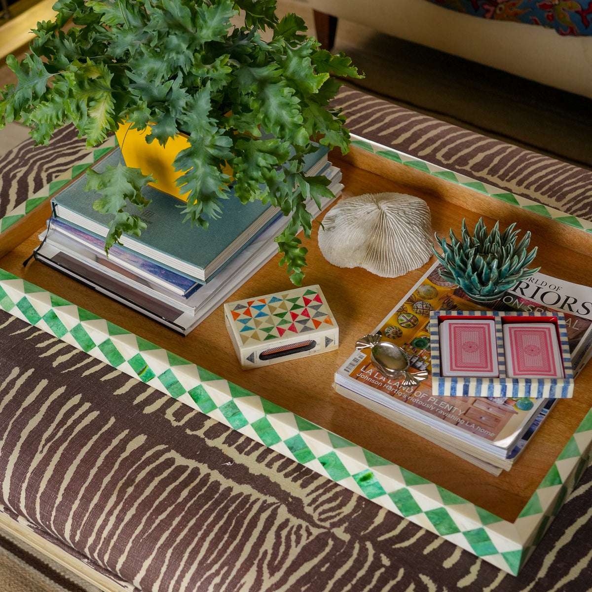 A rectangular wooden serving tray with a green and white checkered border, styled with books, a potted green plant, a white seashell, a small succulent, and a deck of playing cards on a patterned rug.