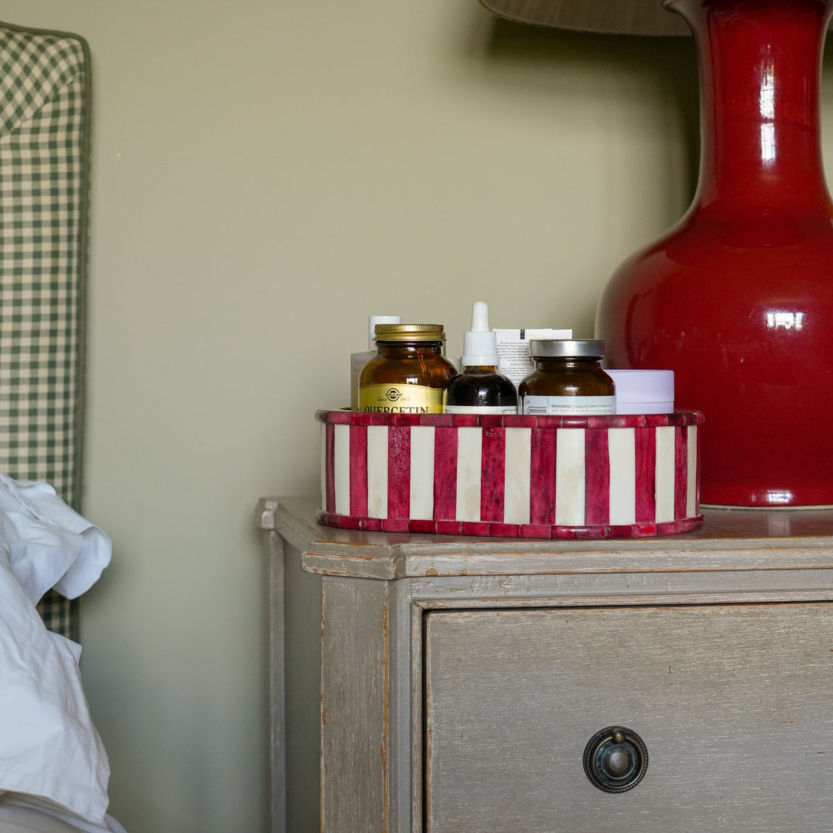 A red and white striped round tray holding small amber bottles and toiletry containers, placed on a rustic gray nightstand beside a red lamp and a bed.