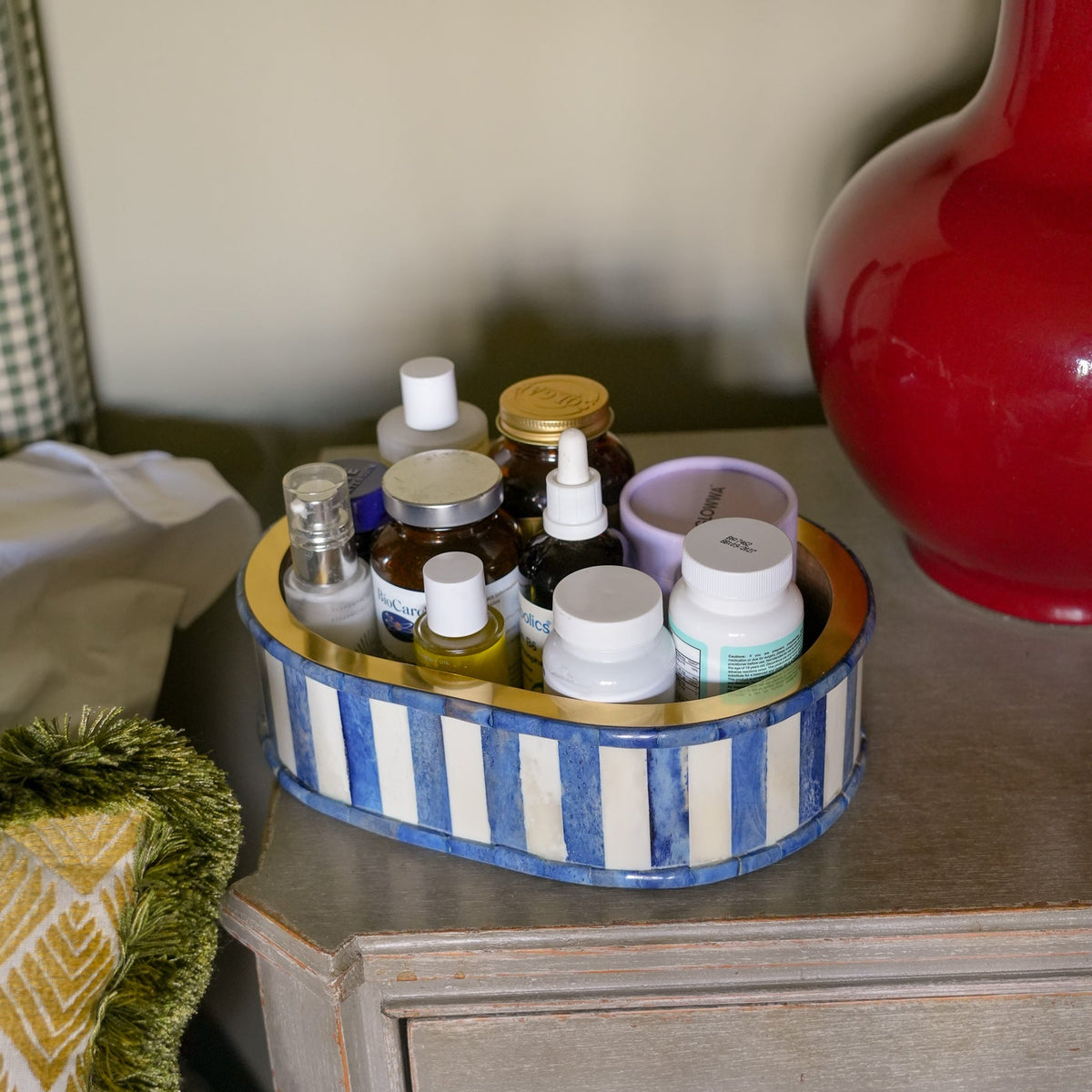 A round, hand-painted tray with blue and white stripes and a gold rim, filled with assorted small bottles and jars, sitting on a distressed gray wooden nightstand next to a red vase.