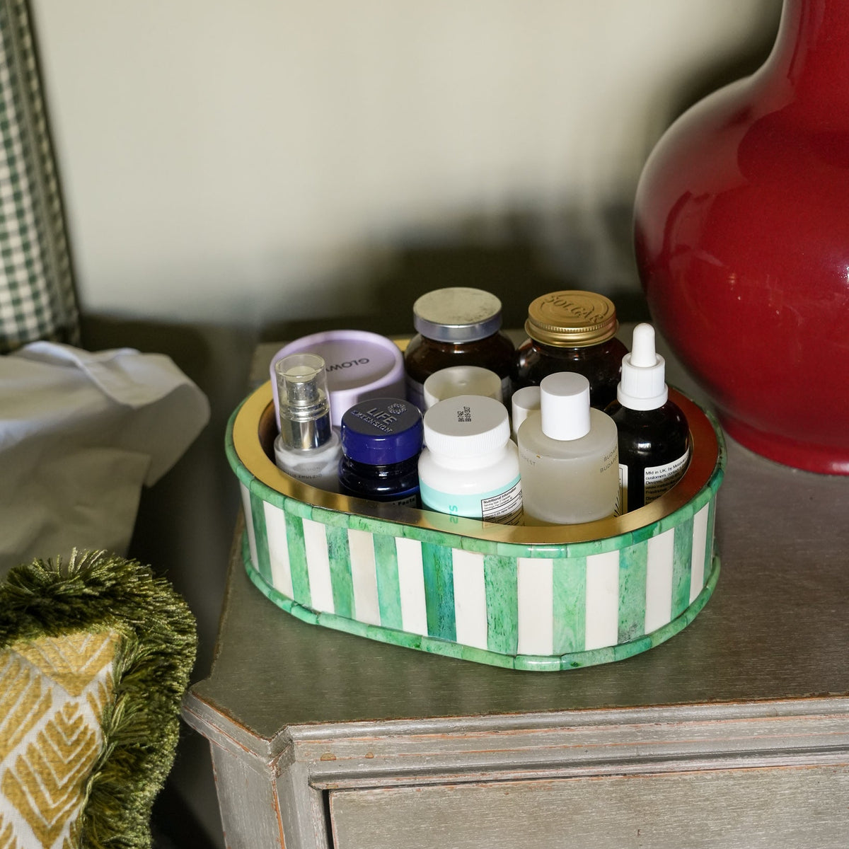 A round green and white striped cosmetic organizer tray holding several small bottles and jars, placed on a wooden bedside table next to a red vase.
