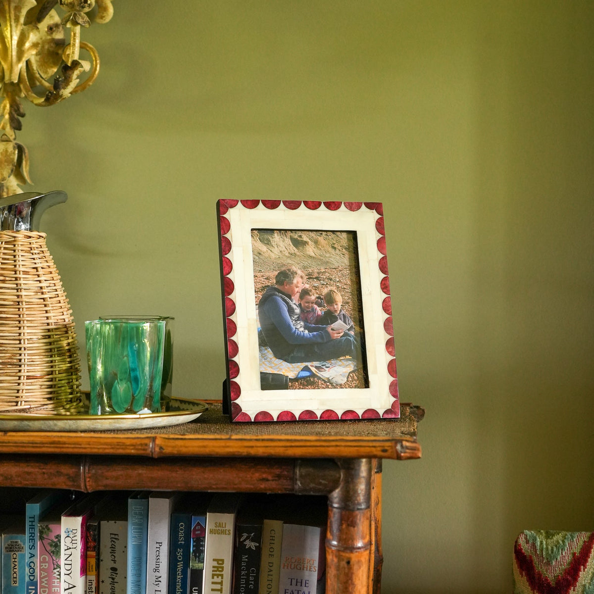 A rectangular picture frame with a red scalloped border and white mat holding a family photo, displayed on a wooden bookshelf against an olive green wall.