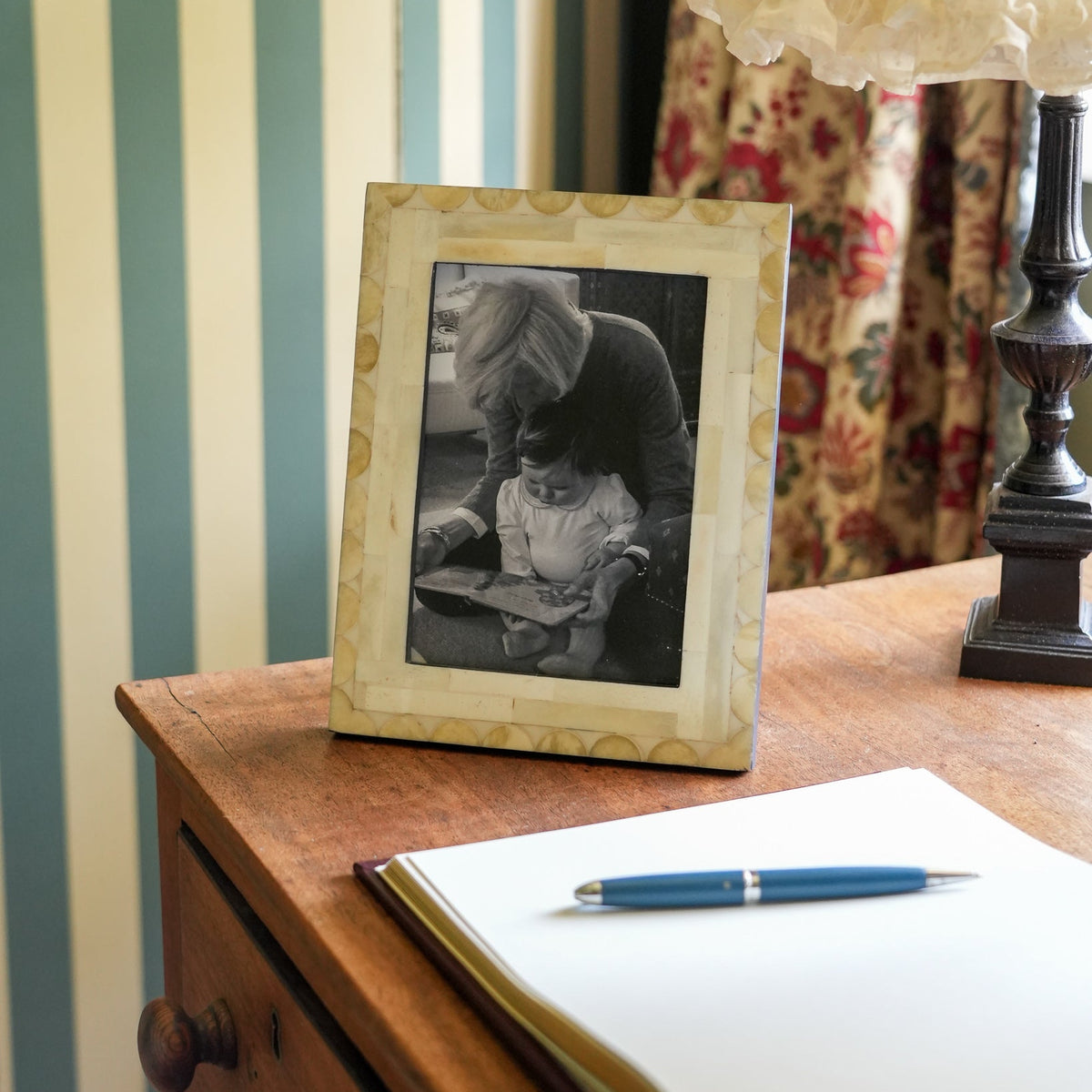 A rectangular picture frame with a decorative cream border holding a black-and-white photo of a woman and child, displayed on a wooden bedside table next to an open notebook and blue pen, with striped wallpaper and a lamp in the background.