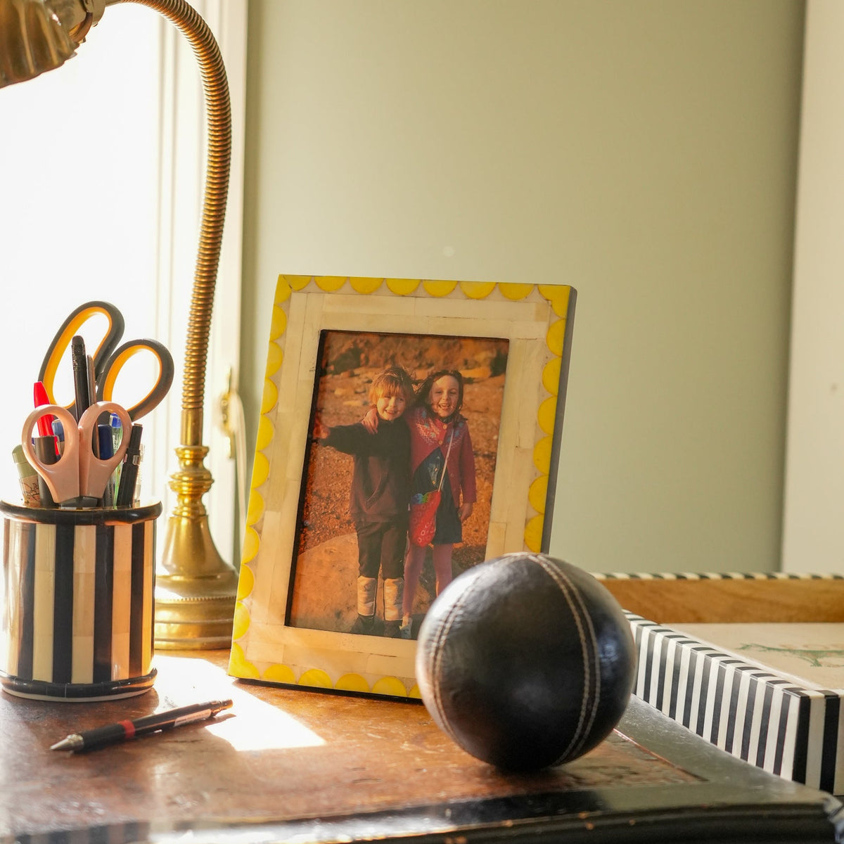 A yellow scalloped picture frame holding a vintage family photograph, styled on a dark wooden desk alongside a brass desk lamp, a striped pencil cup with scissors, and a black leather baseball.