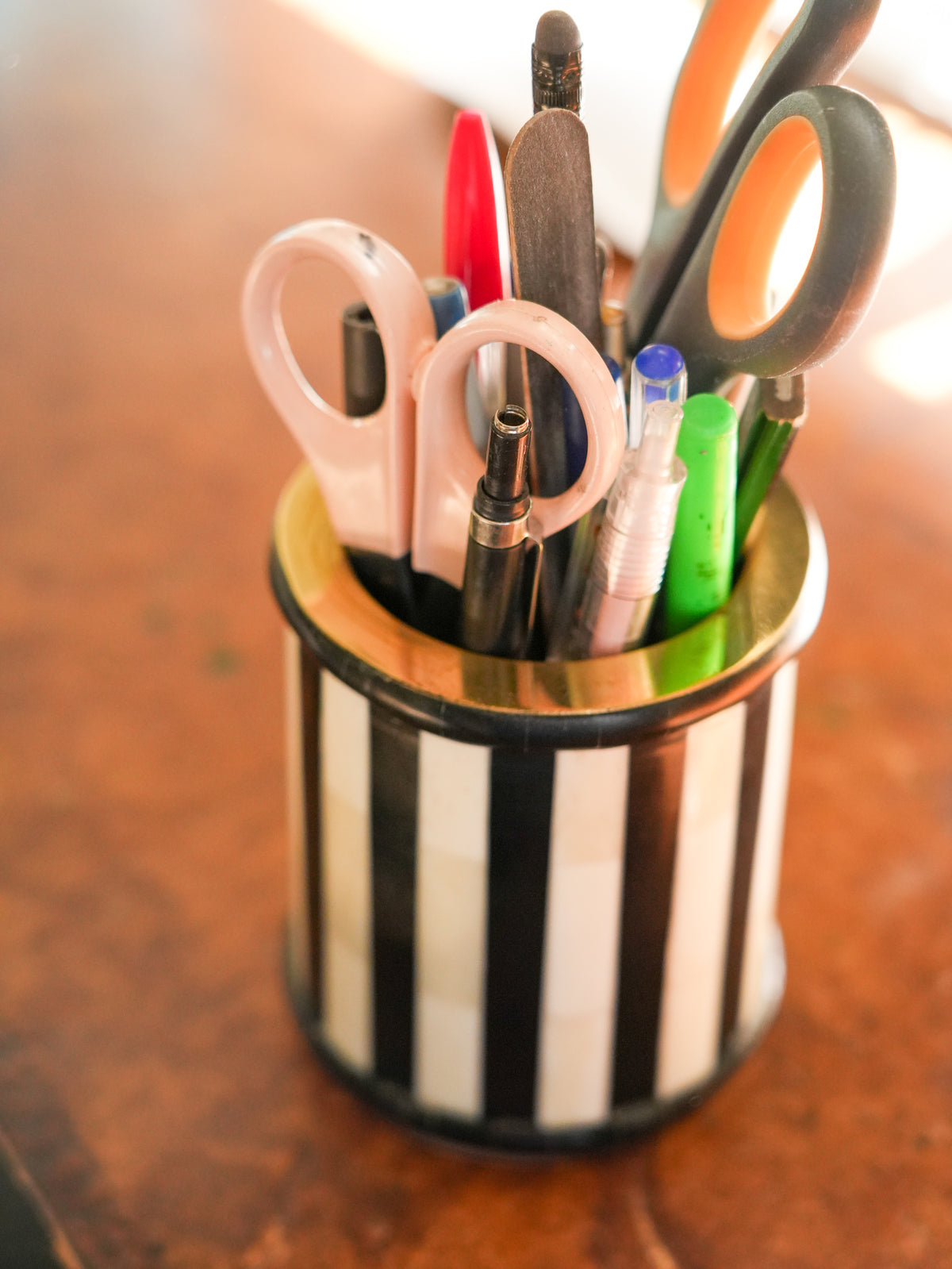 A black and white vertically striped metal pencil cup filled with assorted office supplies, including scissors, pens, and markers, set on a warm brown wooden surface.