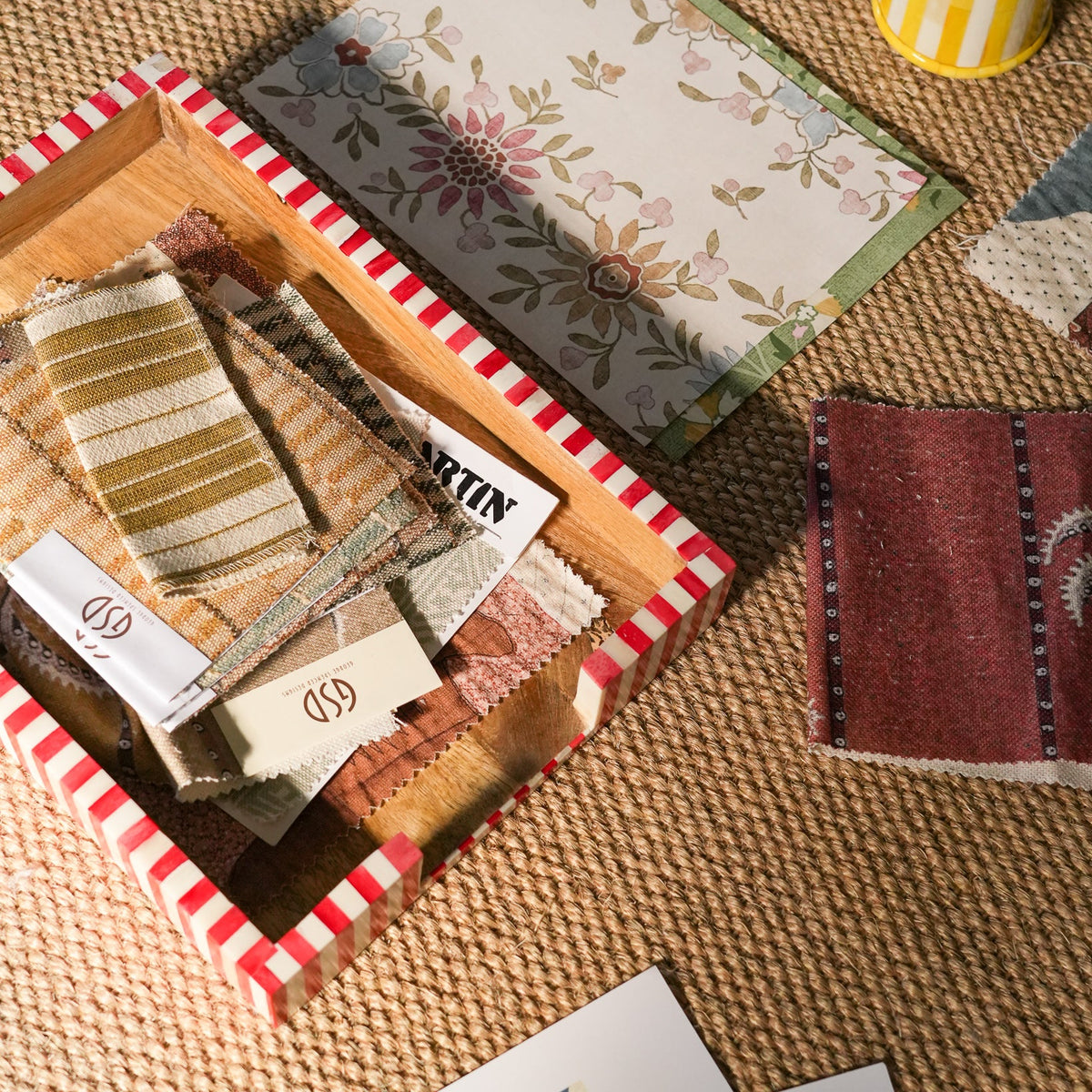 A styled flat-lay of fabric and wallpaper samples arranged on a woven jute rug. A red-and-white striped box holds striped and floral fabric swatches with tags attached, including one tag reading 'MARTIN'.