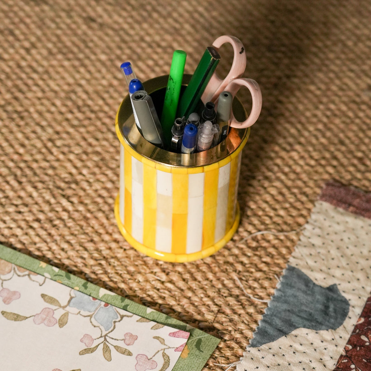 A yellow and white striped metal pencil cup filled with pens, markers, and pink-handled scissors, placed on a woven jute surface with patterned paper pieces nearby.