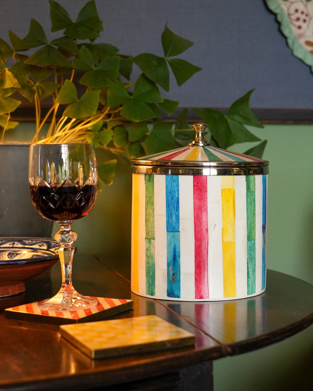 A cylindrical lidded storage jar with vertical multicolor stripes in blue, red, yellow, green, and white, styled on a dark wooden table next to a glass of red wine in a home setting.
