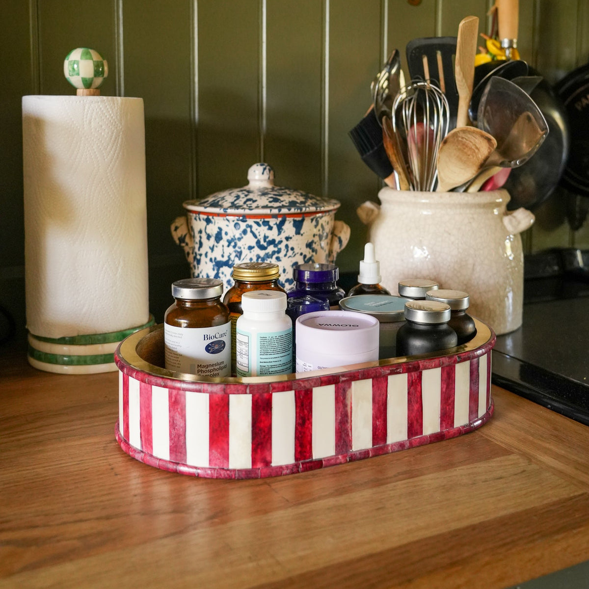 A red and white striped oval tray holding assorted small bottles and jars, placed on a wooden countertop in a kitchen setting with a paper towel holder, ceramic pot, and utensil holder visible.