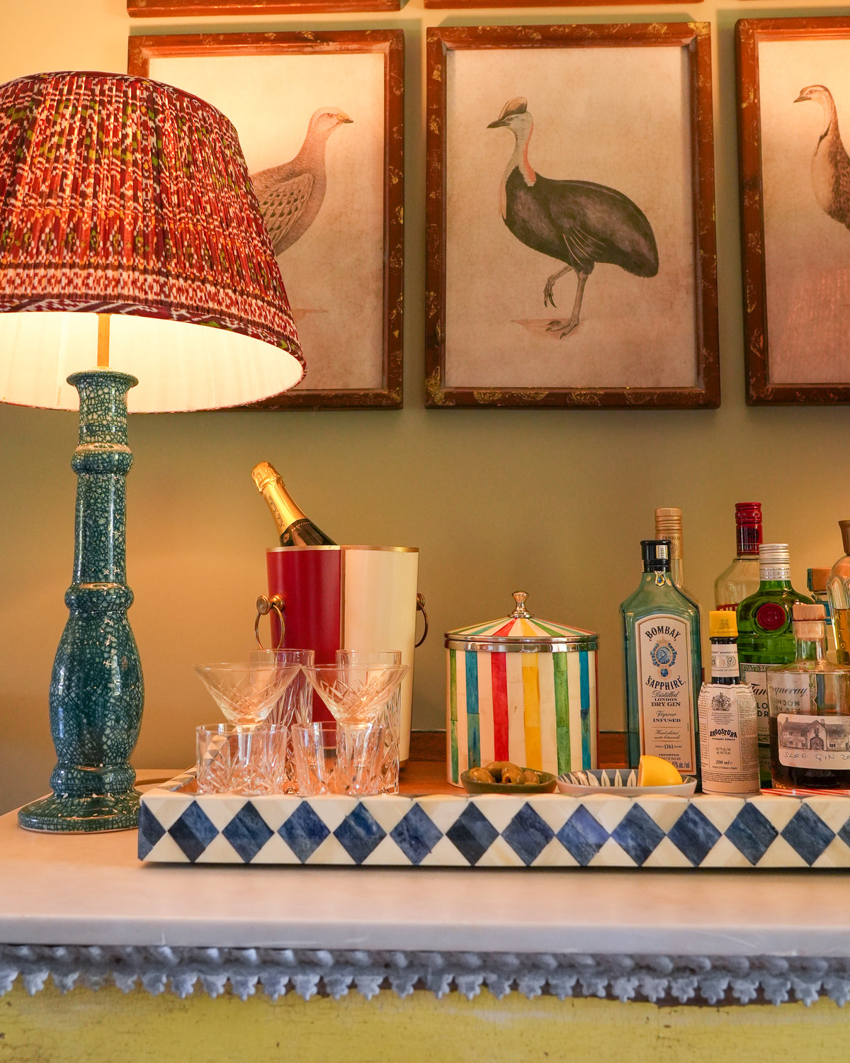 A styled bar counter scene with a blue-green ceramic table lamp, a red patterned lampshade, crystal glasses, a champagne bucket, colorful bottles, and framed bird artwork on the wall behind.