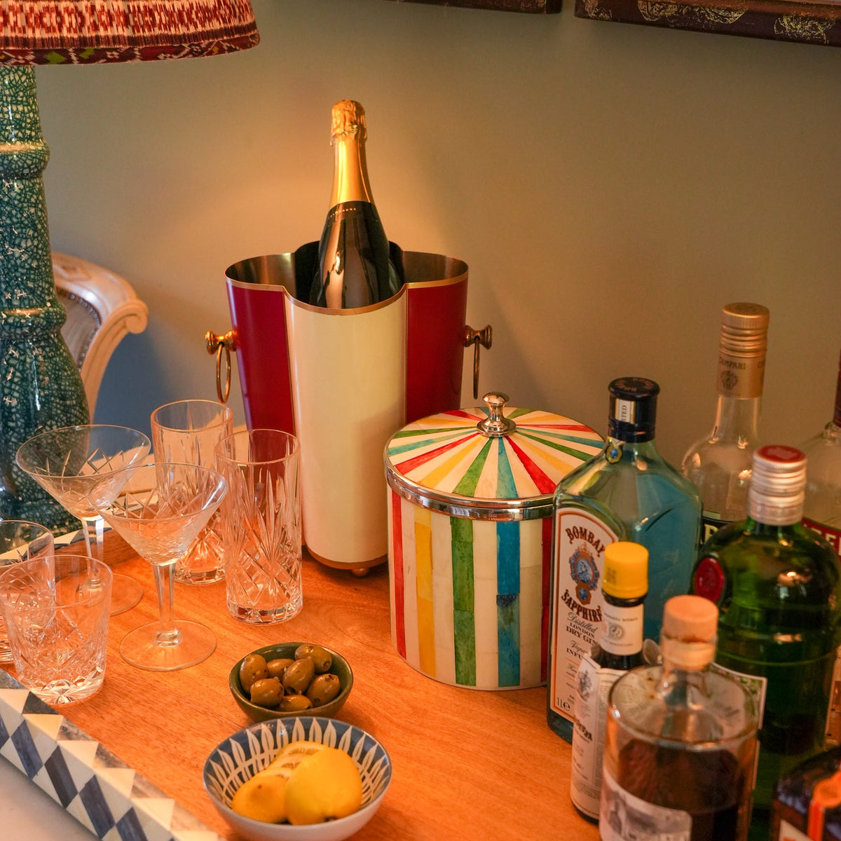 A styled bar or cocktail station with assorted bottles, glasses, olives, lemons, and a striped lidded container on a wooden table, with a champagne bucket and framed wall art in the background.