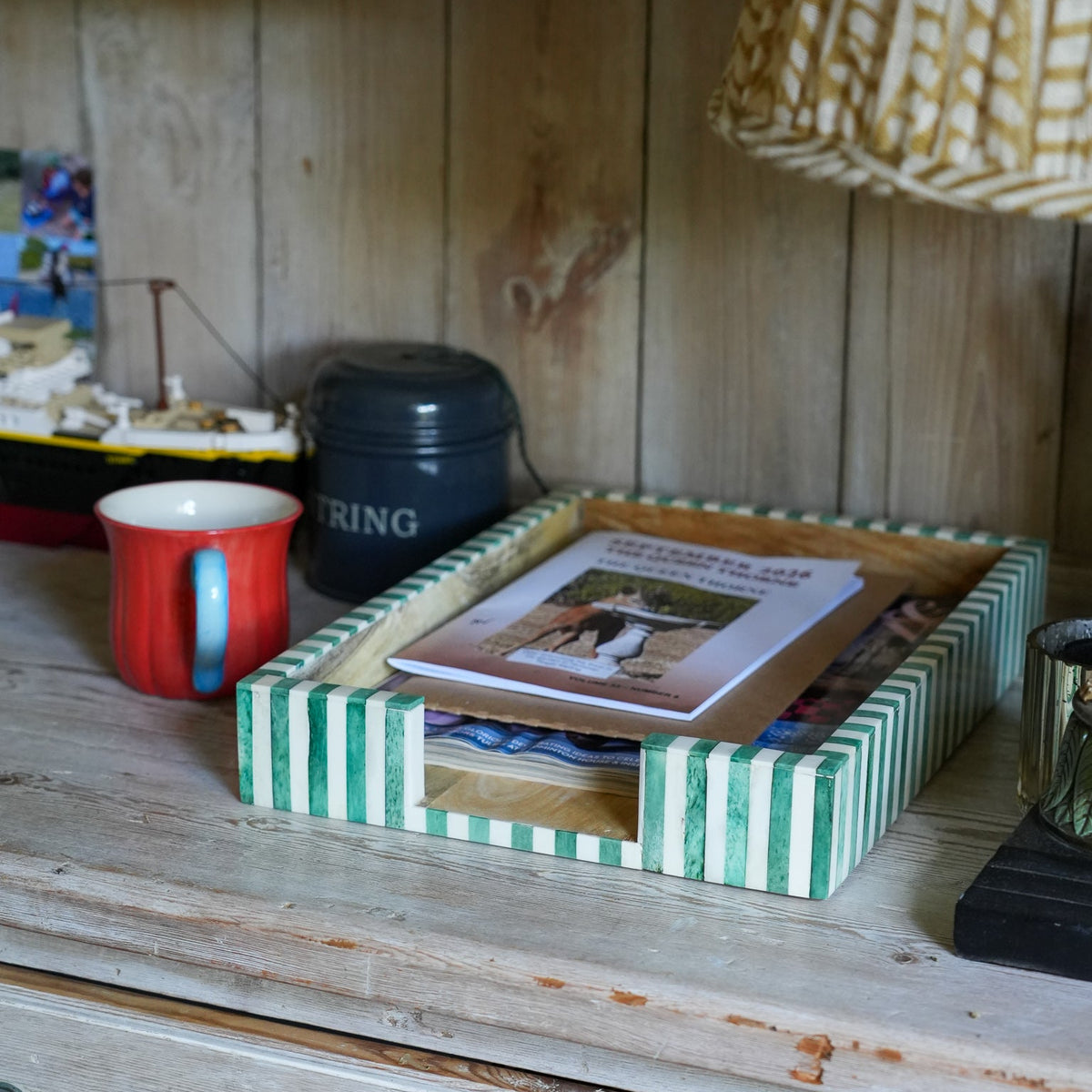 A green and white striped wooden tray holding a magazine, set on a rustic wooden dresser next to a red mug in a home interior setting.