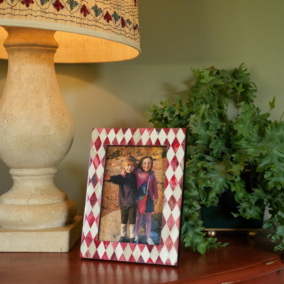 A diamond-patterned picture frame with red and white argyle and a floral border, holding a photo of two children, styled on a wooden table next to a lamp and a green plant.