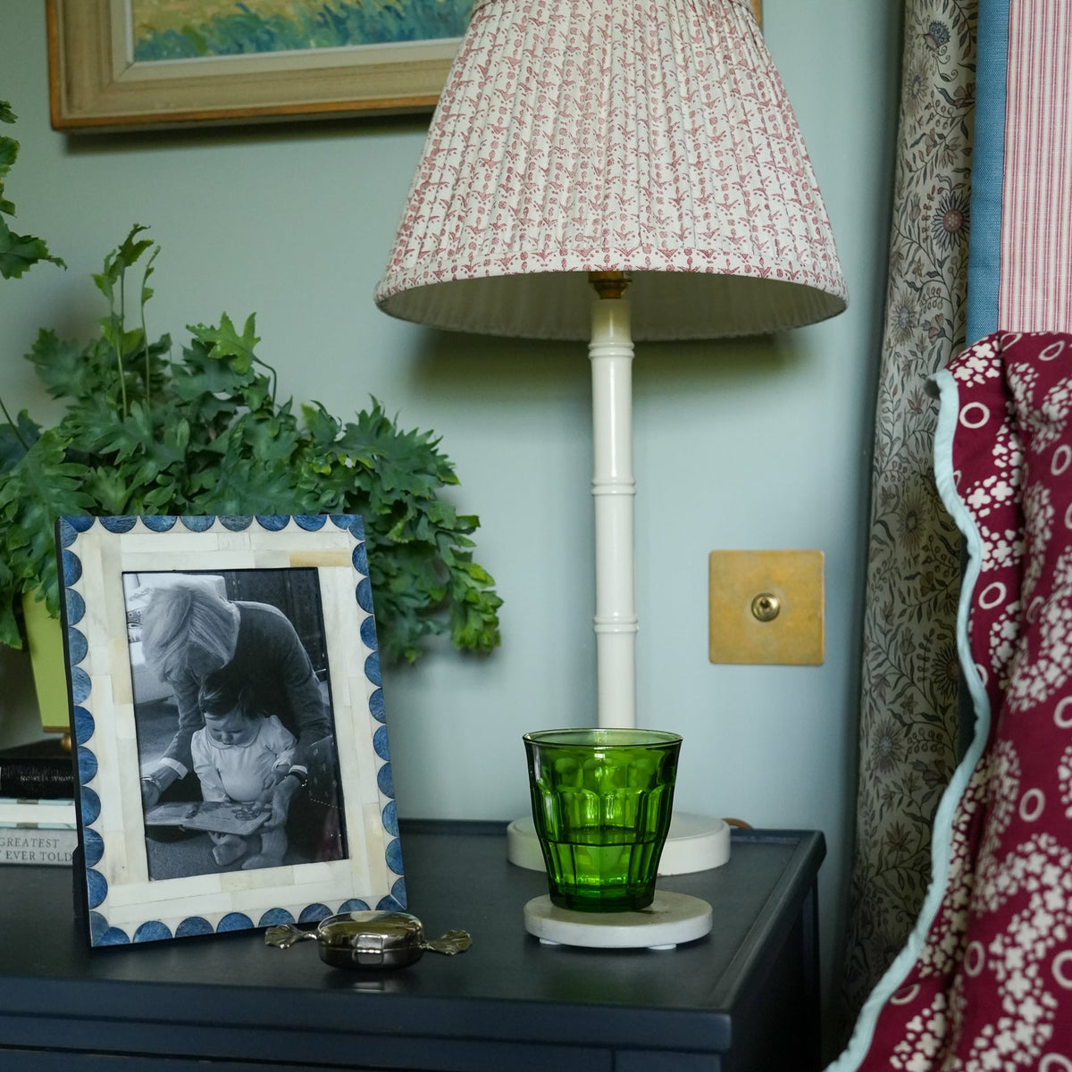 A vintage-style table lamp with a textured pink and white patterned shade and a white bamboo-look base, styled on a dark blue bedside table next to a green glass cup, a framed black-and-white photo, and a blue wall.