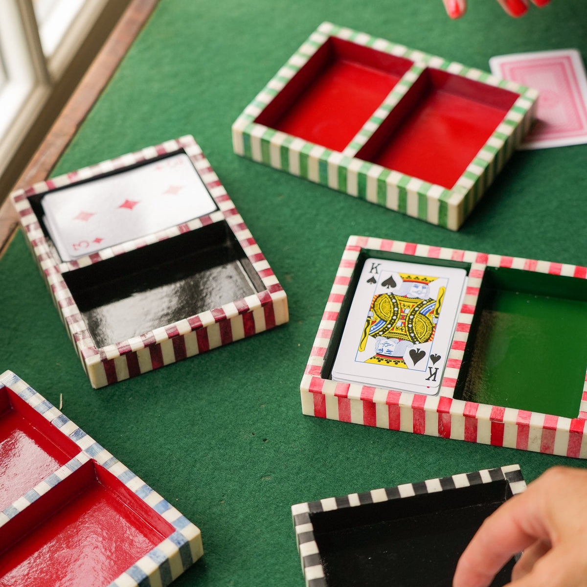 Several striped card boxes with colorful interior lids are arranged on a green felt table, with hands reaching in to lift the lids and reveal playing cards.