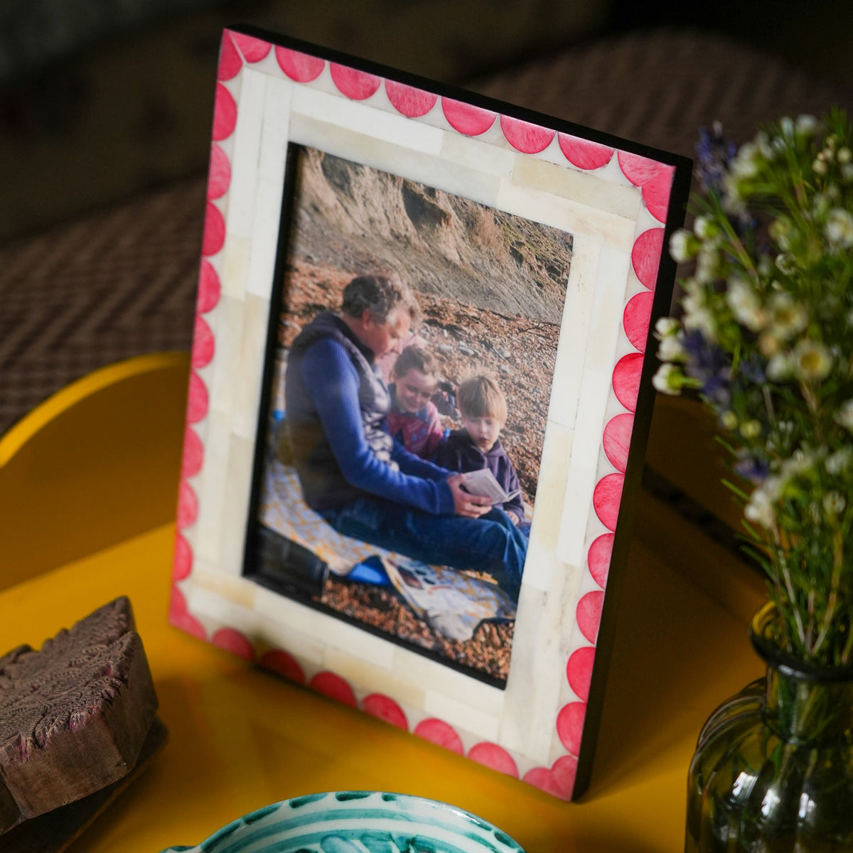 A rectangular picture frame with a red scalloped border and a cream center mat, holding a photo of a man and two children. The frame is styled on a yellow tray next to a green glass vase with flowers and a decorative teal ceramic dish.