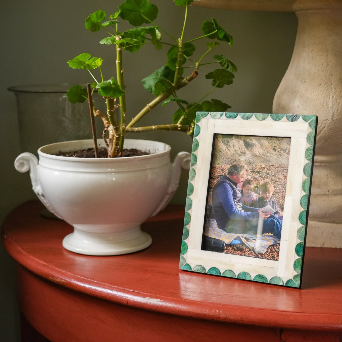 A rectangular photo frame with a green scalloped border stands on a reddish-brown wooden table next to a white planter with green leaves. The frame contains a photograph of a man and child.