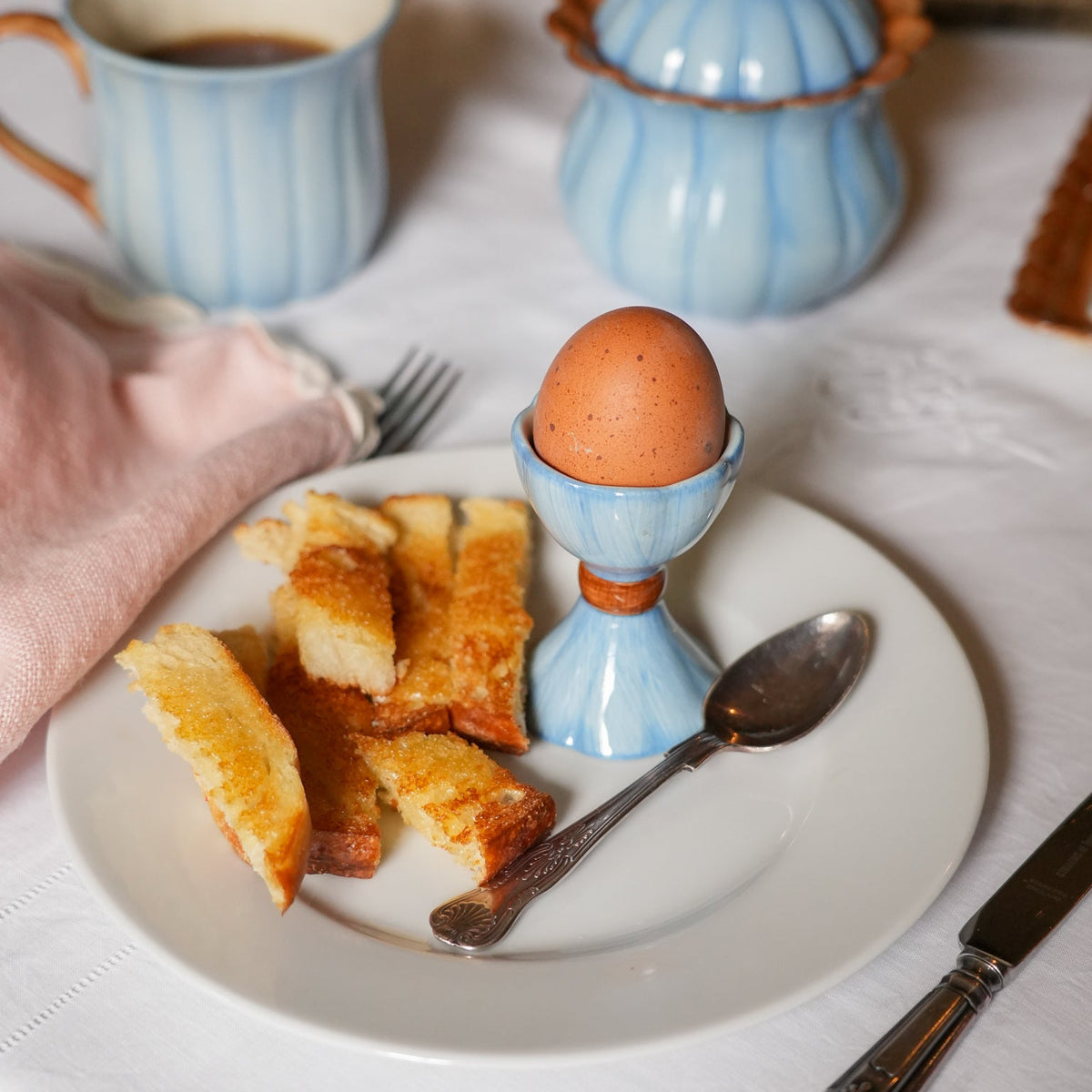 A breakfast table setting featuring a blue ribbed egg cup holding a brown speckled egg, accompanied by toast on a white plate, a blue mug, and a blue lidded jar in the background.