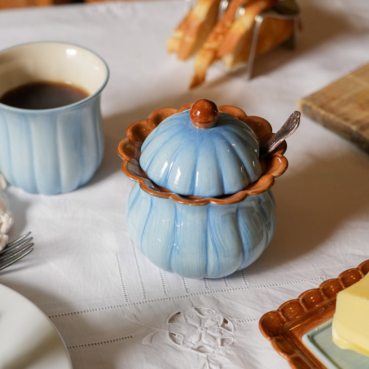A light blue ribbed ceramic sugar bowl with a matching lid and spoon, set on a white tablecloth alongside a matching blue cup of coffee and bread on a table.