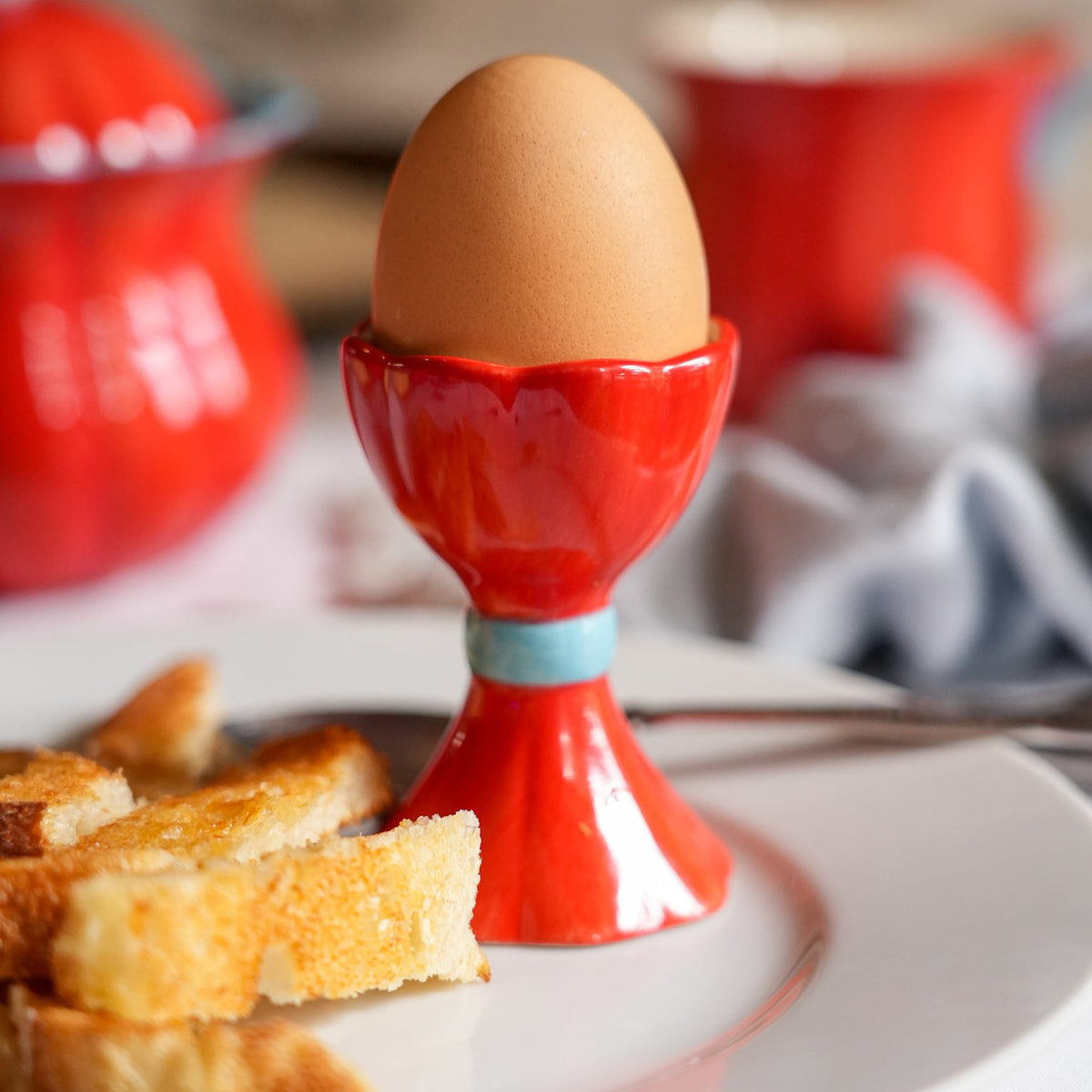 A brown egg sits in a glossy red ceramic egg cup with a light blue band, set on a white plate with toasted bread in the foreground and a red teapot in the soft-focus background.