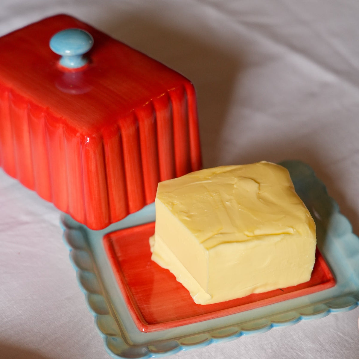 A butter dish set on a white tablecloth, featuring a bright red ribbed butter box with a light blue knob and a matching scalloped light blue dish holding a square of pale yellow butter.