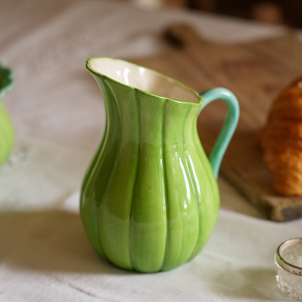 A green ribbed ceramic pitcher with a turquoise handle, set on a white tablecloth with a blurred rustic background.
