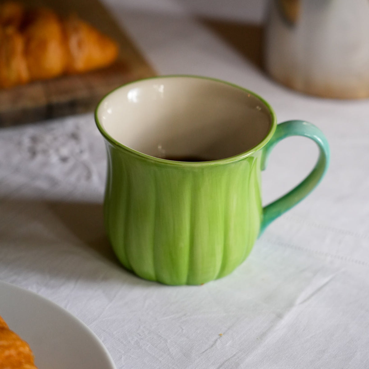 A green ceramic mug with a ribbed texture and a teal handle, filled with coffee on a white tablecloth with a blurred croissant in the background.