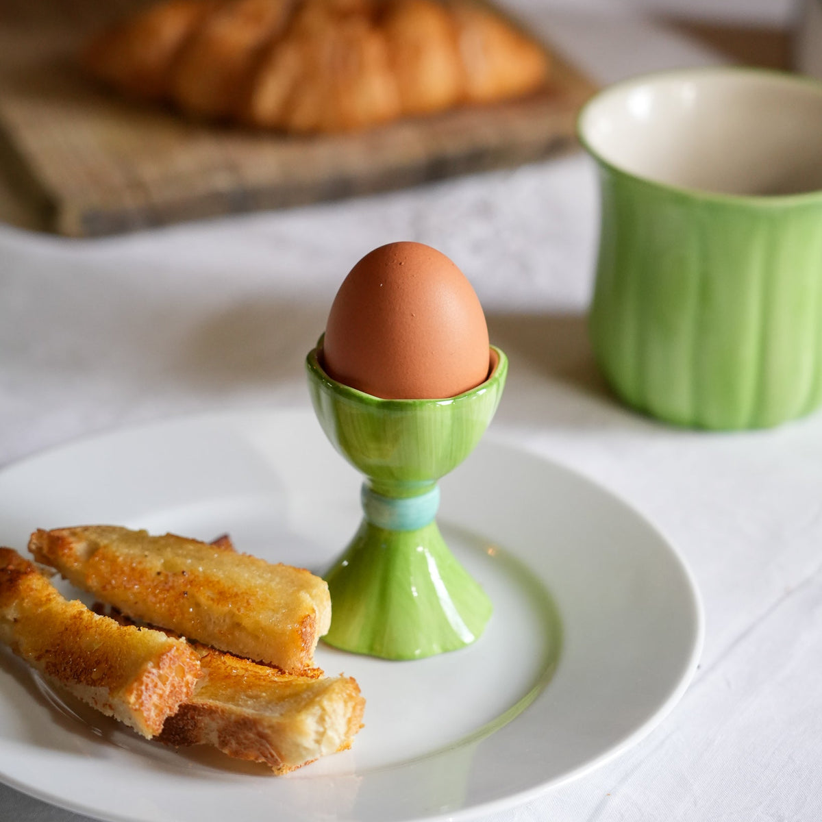 A green ceramic egg cup holding a brown egg, set on a white plate with toast in the foreground and a green mug in the background on a white tablecloth.