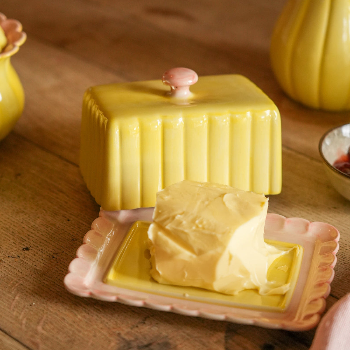 A yellow ceramic butter dish with a pink knob and a pink scalloped plate holding a slice of butter, set on a rustic wooden table.