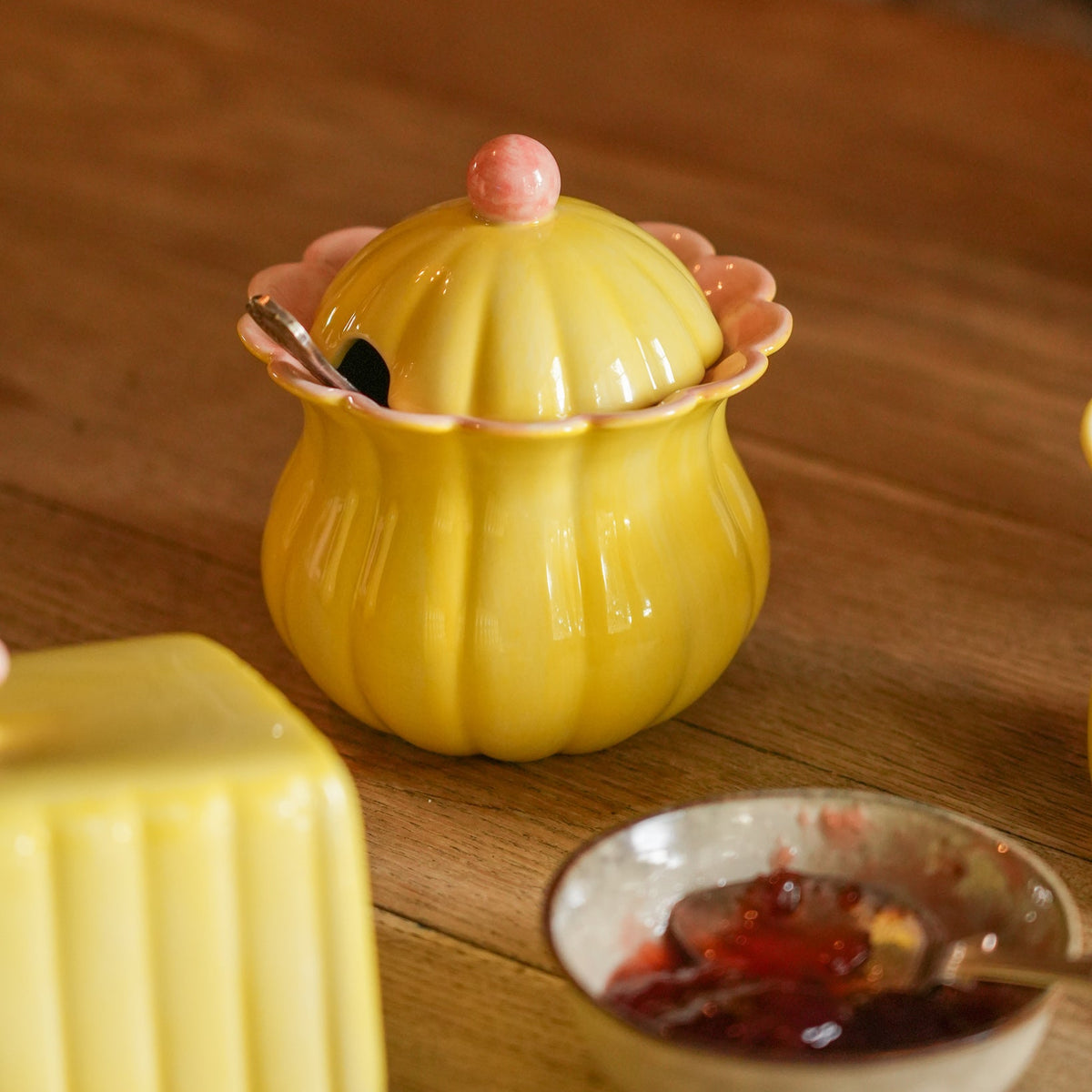 A yellow ribbed ceramic jar with a pink lid knob and scalloped pink rim, shown on a wooden table with butter and jam nearby.