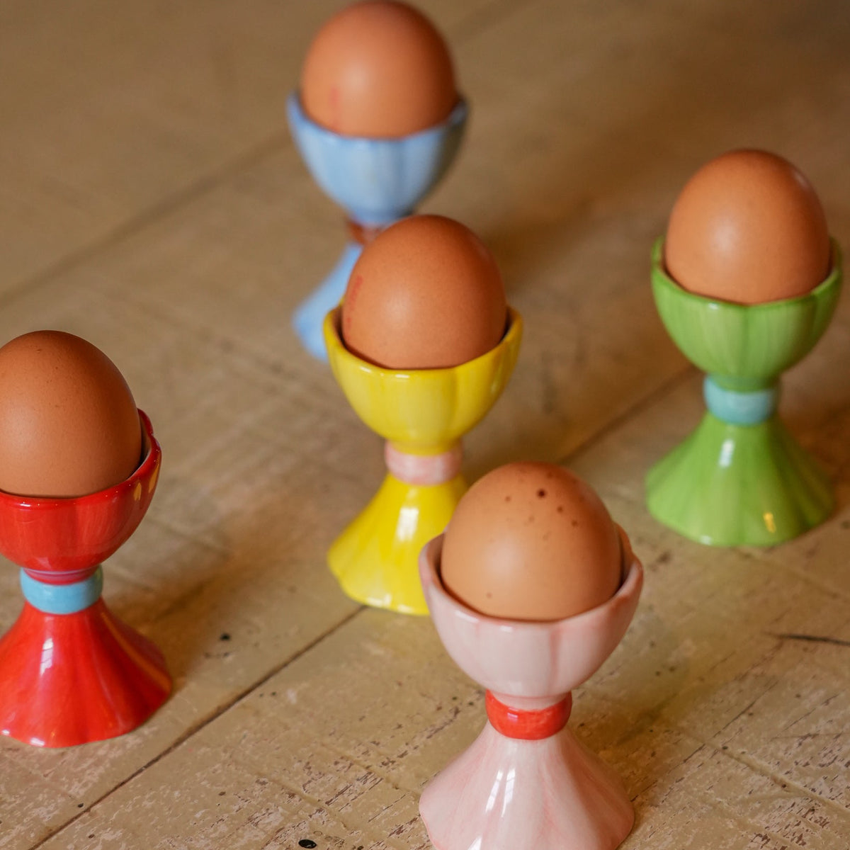 Five small ceramic egg cups in red, yellow, green, blue, and pink, each holding an brown egg, arranged on a rustic wooden tabletop.