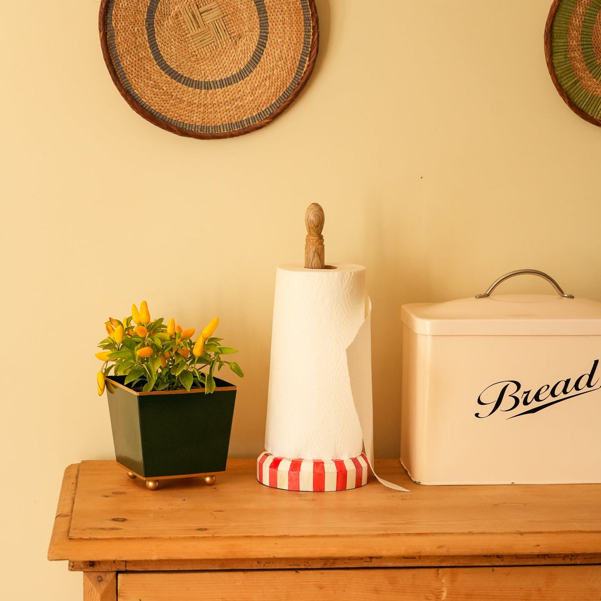 A styled interior scene showing a wooden dresser against a warm cream wall, with a potted yellow flower, a paper towel holder, and a cream bread tin labeled 'Bread'.