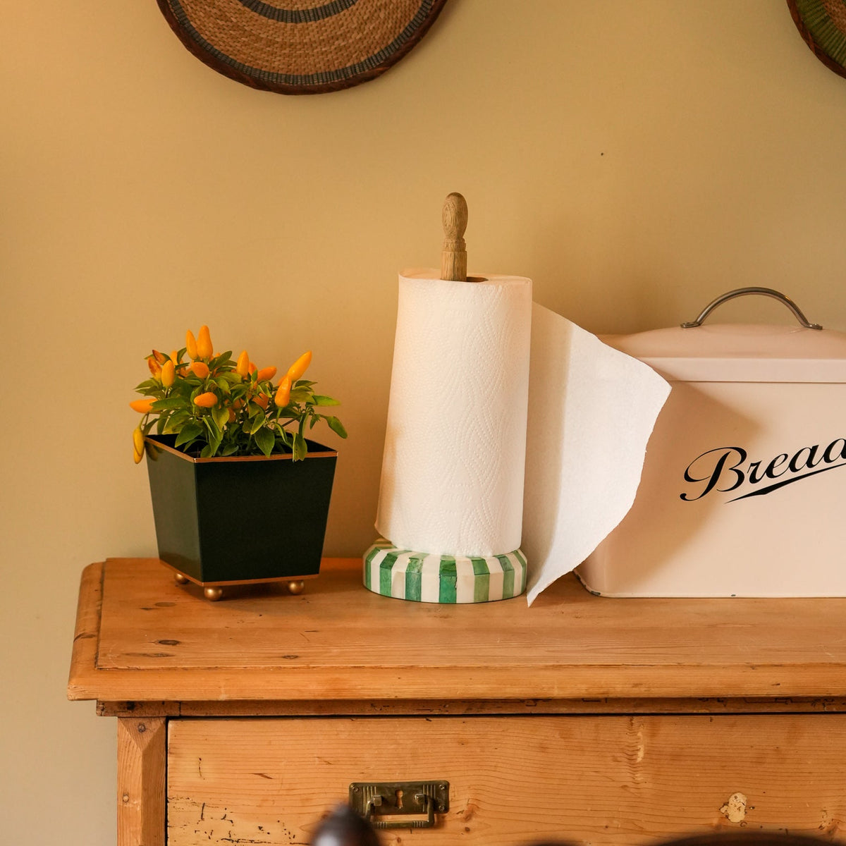 A kitchen countertop scene with a paper towel roll on a green-and-white striped holder, a small potted orange flower, and a cream bread box with partial script lettering, set on a wooden cabinet against a warm beige wall.