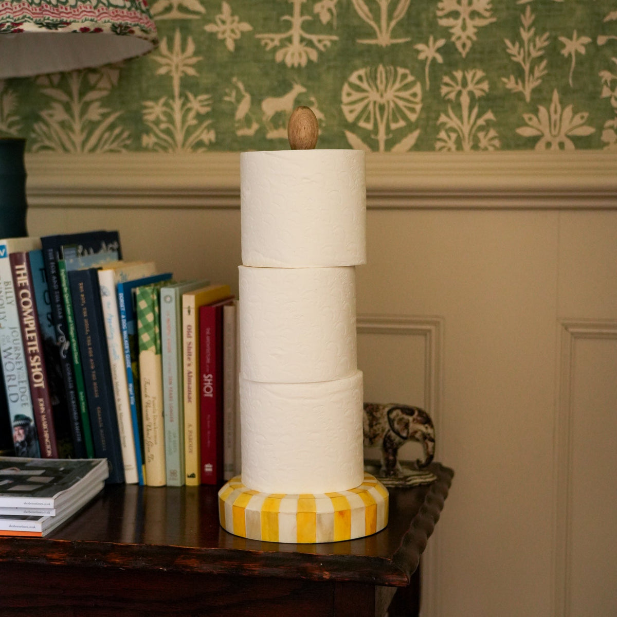 A stack of four white toilet paper rolls on a yellow and white striped holder with a small wooden top, displayed on a dark wooden table in a home setting with books and patterned wallpaper in the background.