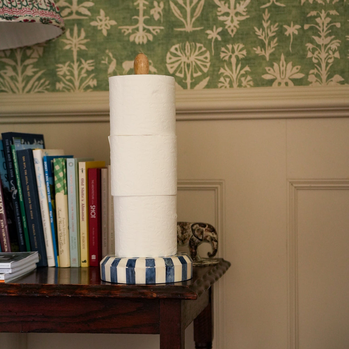 A stack of white paper towels on a striped blue and white holder with a wooden handle, set on a dark wooden table against a green and beige patterned wallpaper background.