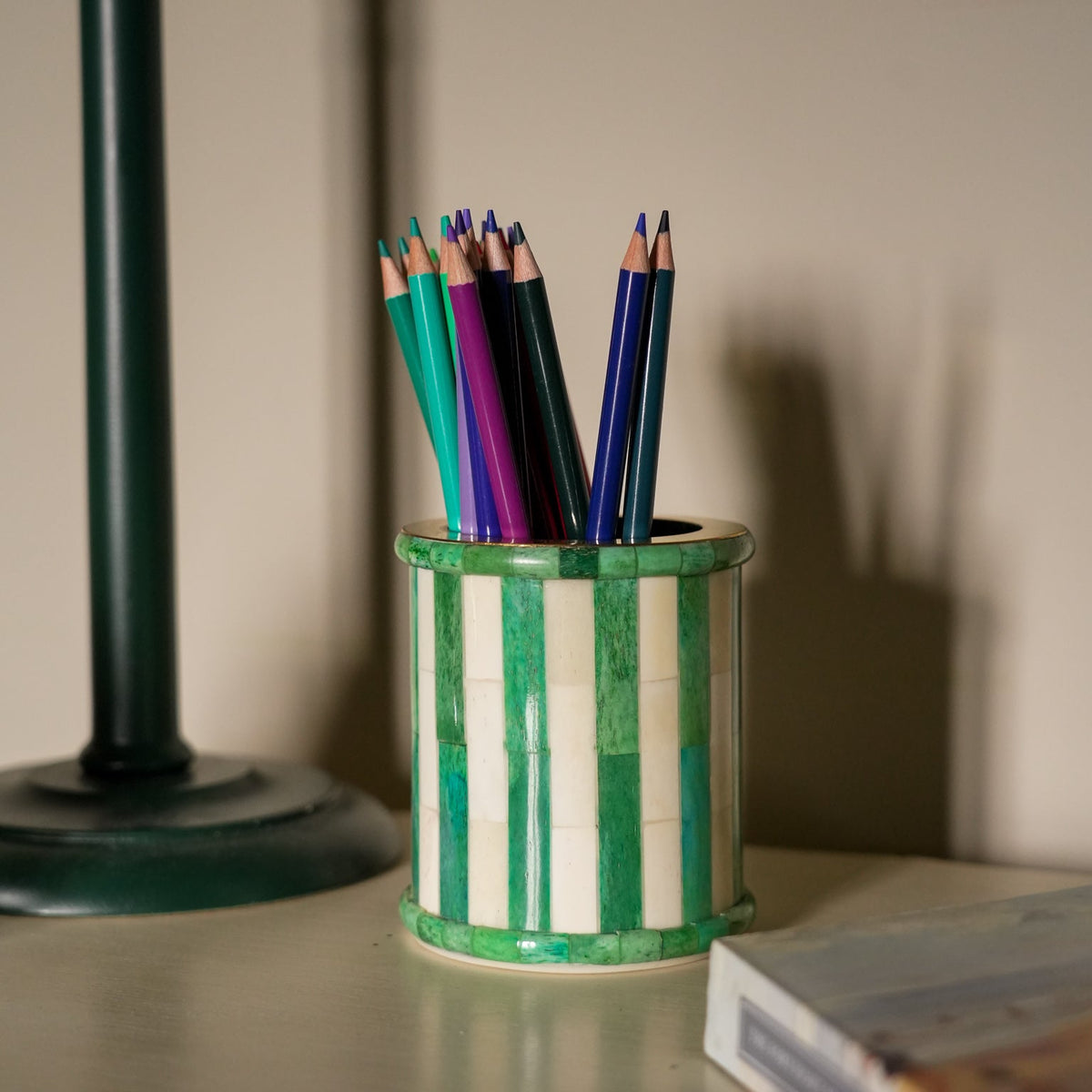 A cylindrical ceramic pencil holder with vertical green and white stripes, filled with colored pencils, sitting on a tabletop next to a dark green lamp base.