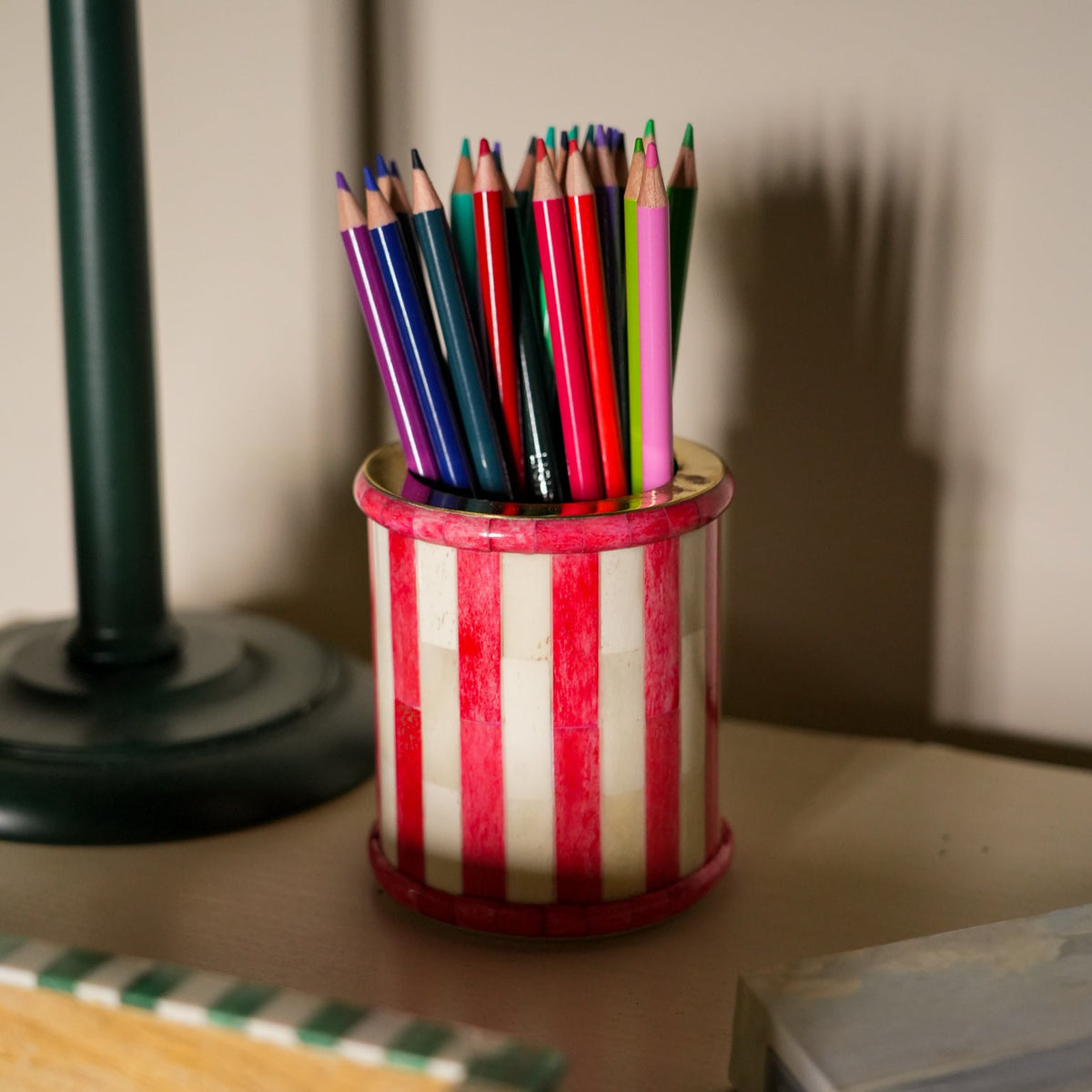 A red and white vertically striped pencil holder filled with colored pencils, sitting on a tabletop next to a dark green lamp base.