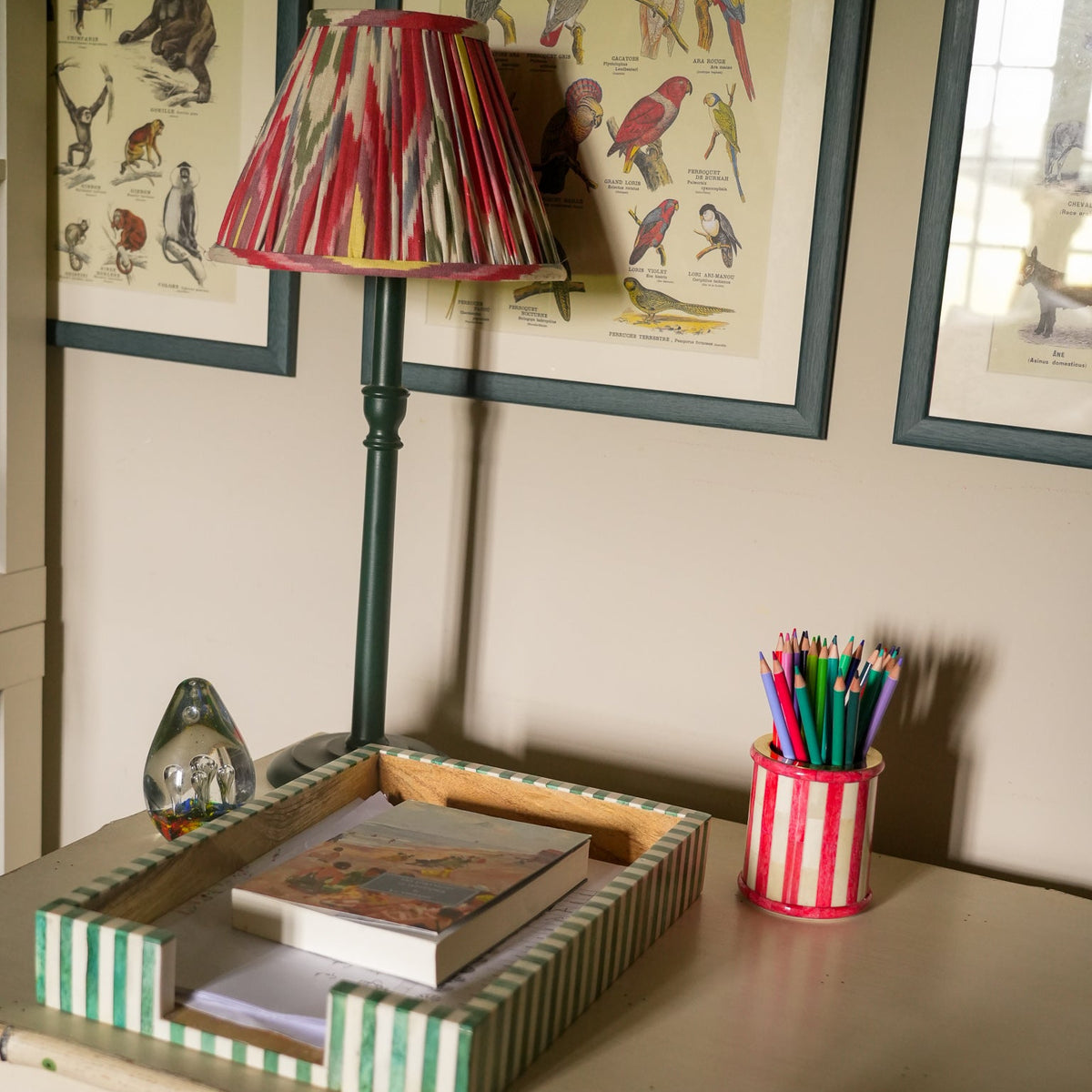 A styled home office corner featuring a green desk with a striped green-and-white tray, a striped pencil cup, a striped lamp, and framed bird illustrations on the wall.