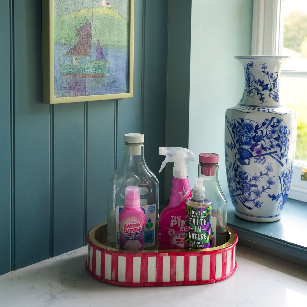 A styled interior corner with teal paneled walls, a brass wall lamp, a framed boat painting, and a large blue-and-white ceramic vase on a windowsill. In front of the window sits a pink-and-white striped tray holding several cleaning spray bottles.