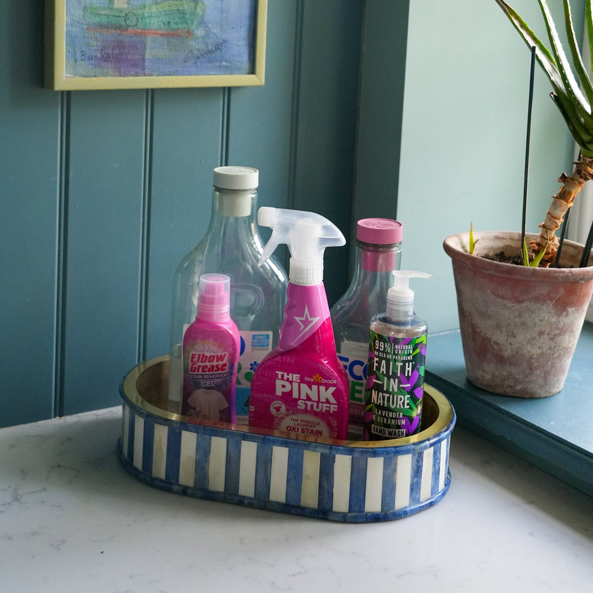 A set of cleaning products arranged in a blue and white striped oval tray on a countertop, with a teal paneled wall, framed artwork, and a potted plant in the background.