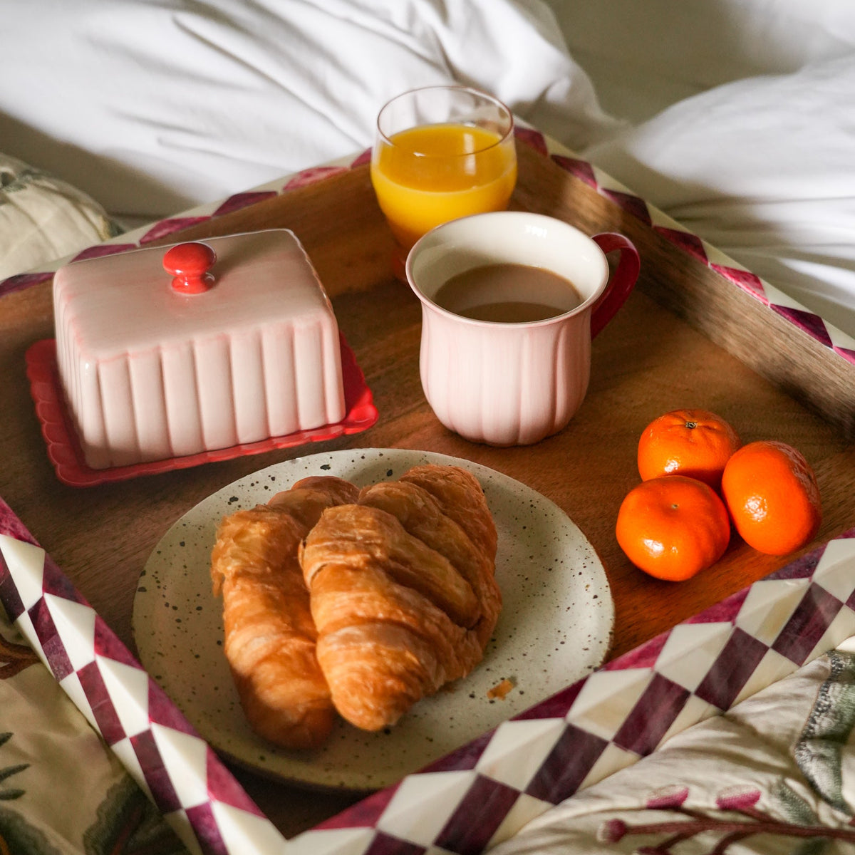 A breakfast tray set on white bedding, featuring croissants on a speckled plate, a pink butter dish, a cup of coffee, a glass of orange juice, and three oranges, all arranged on a patterned checkered blanket.