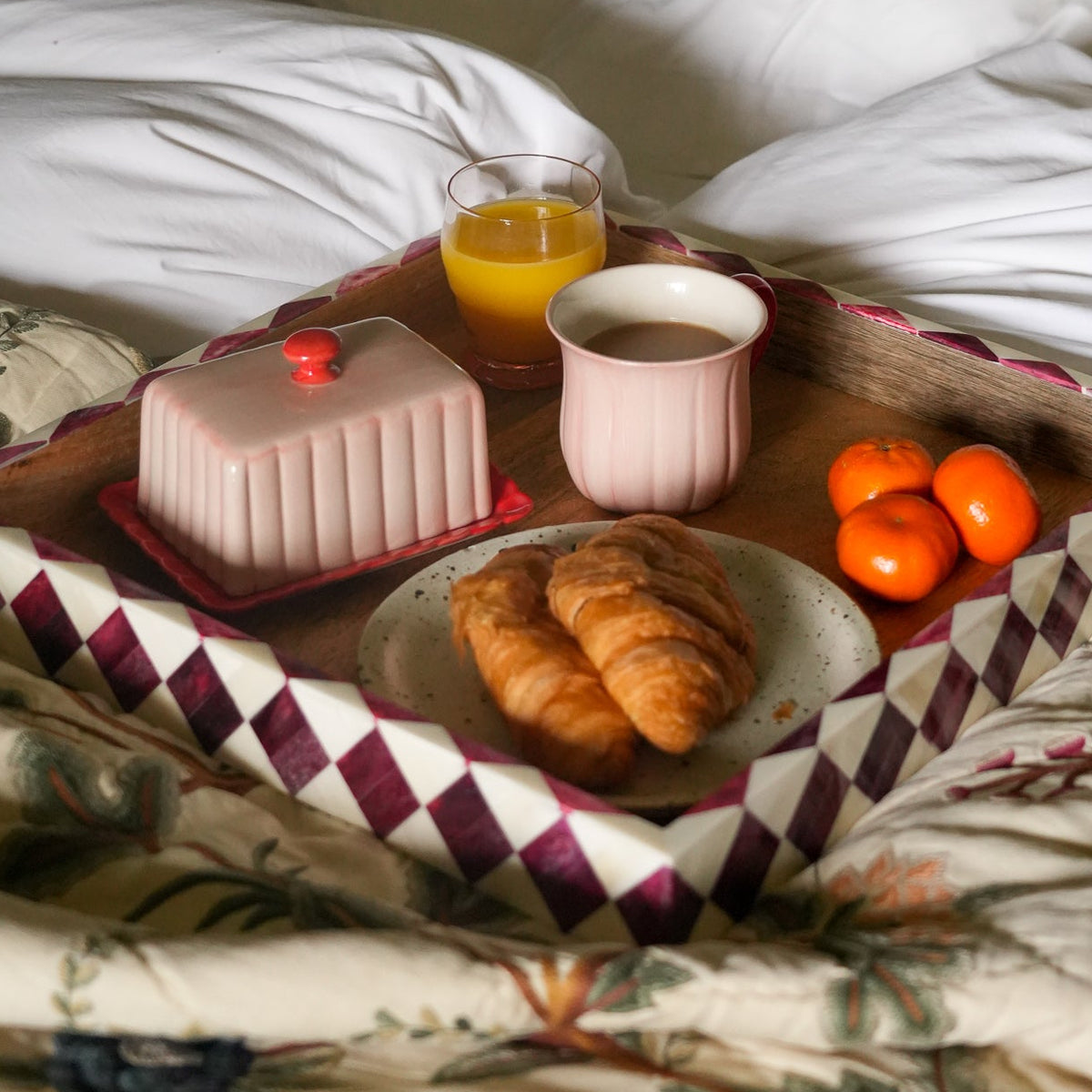 A breakfast tray set on rumpled white bedding, featuring croissants, orange tangerines, a glass of orange juice, a pink cup, and a pink pastry on a patterned tray.