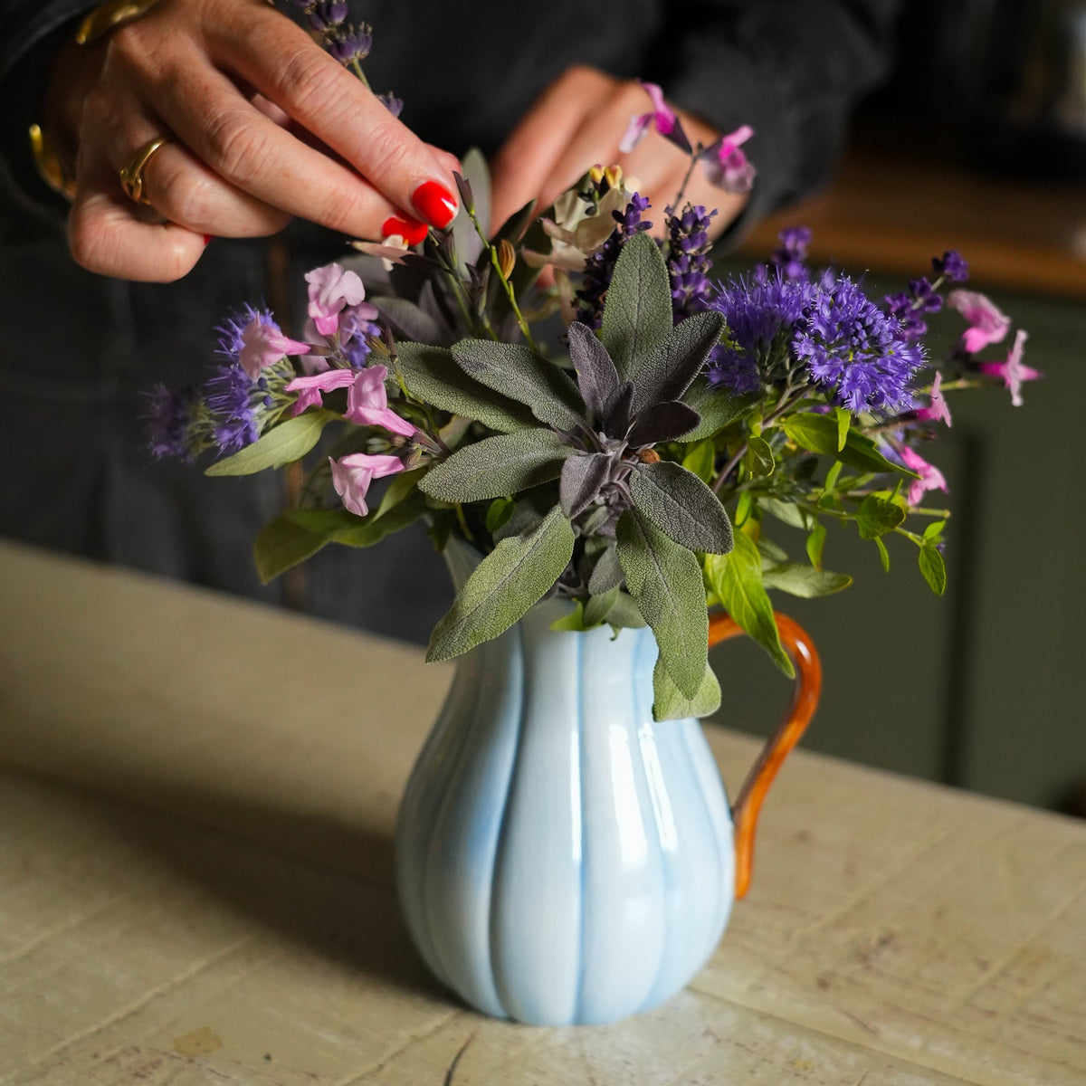 A light blue ribbed ceramic pitcher vase with an orange handle, filled with a bouquet of purple flowers and green leaves, sitting on a rustic wooden table while a person's hands adjust the top of the arrangement.