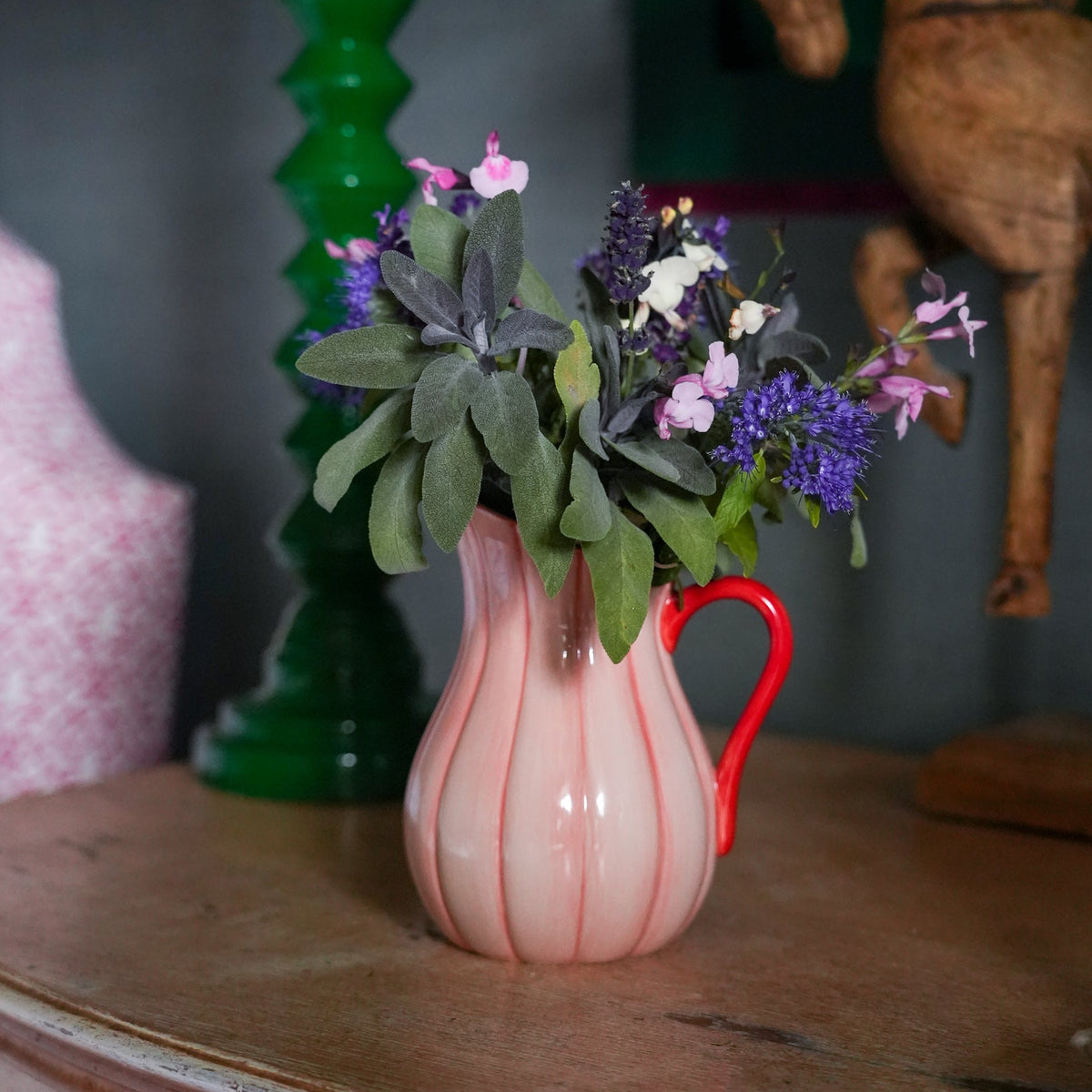 A pink ribbed ceramic pitcher vase with a red handle, filled with green foliage and purple flowers, set on a wooden table in a styled interior.