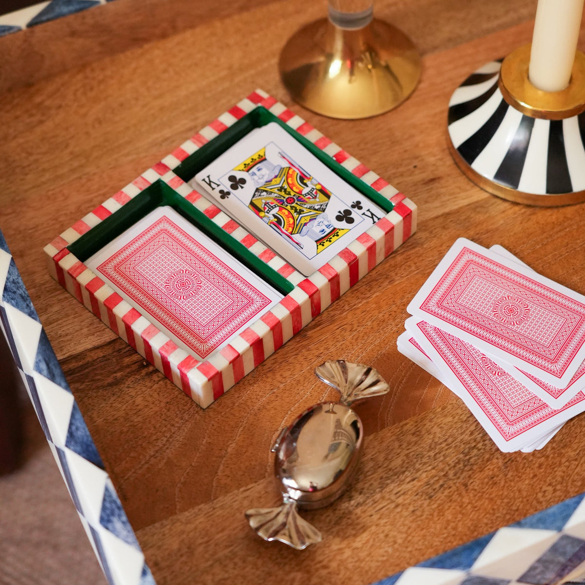 A styled tabletop scene showing a small red-and-white striped box containing playing cards, with additional cards and a wrapped candy on a wooden tray. A candlestick and patterned chair are visible in the background.