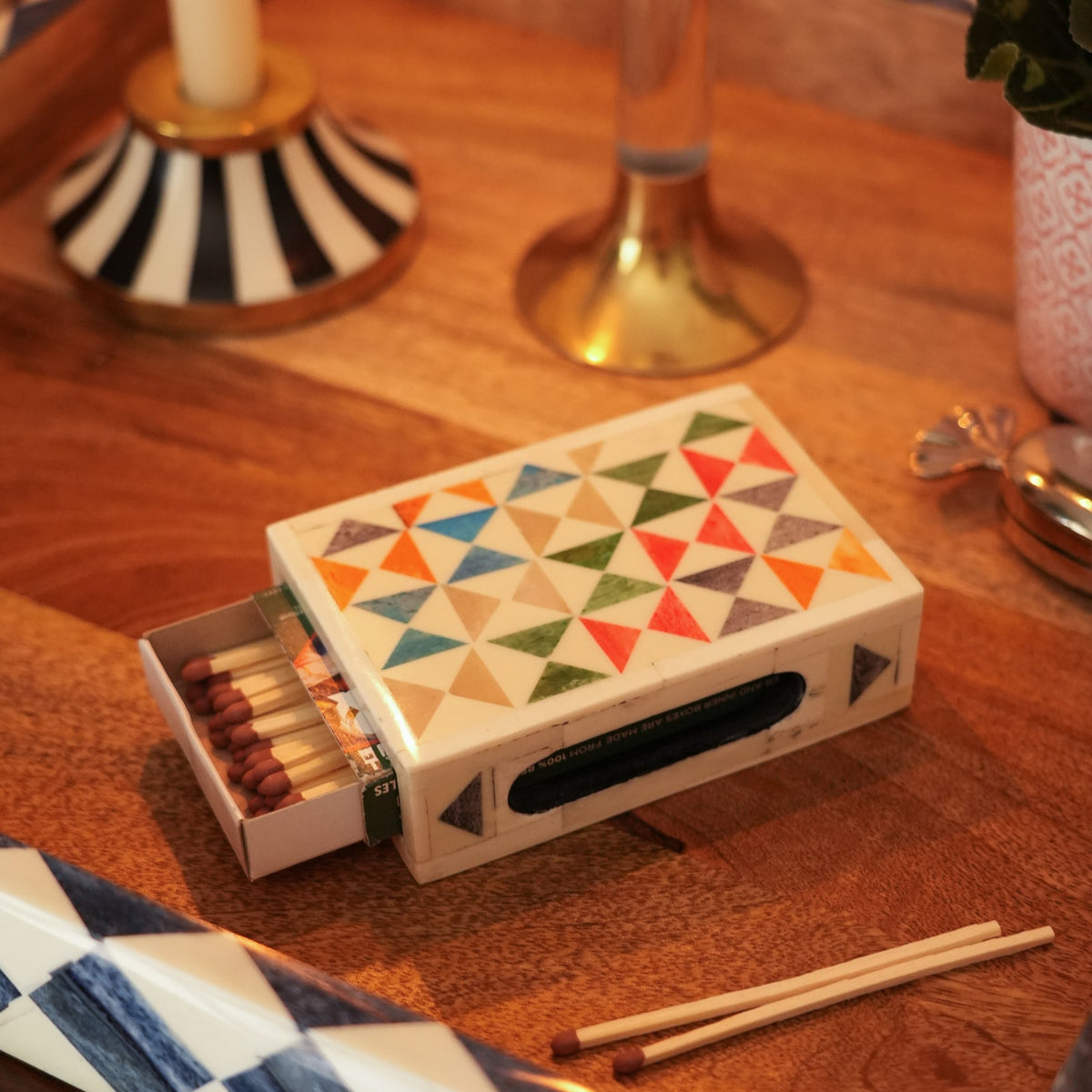 A decorative matchbox with a geometric triangle pattern in multiple colors, shown open with matches inside on a wooden table. The matchbox has a black handle slot and is styled in a warm, home-like setting with other tabletop objects in the background.
